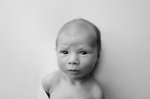 Black and white close-up portrait of a wide-eyed newborn baby with a curious, furrowed expression against a soft, plain background.