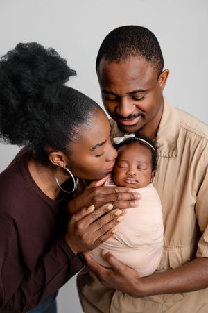 Smiling parents tenderly kiss and cradle their sleeping newborn baby wrapped in a soft blush blanket in a bright newborn family portrait.