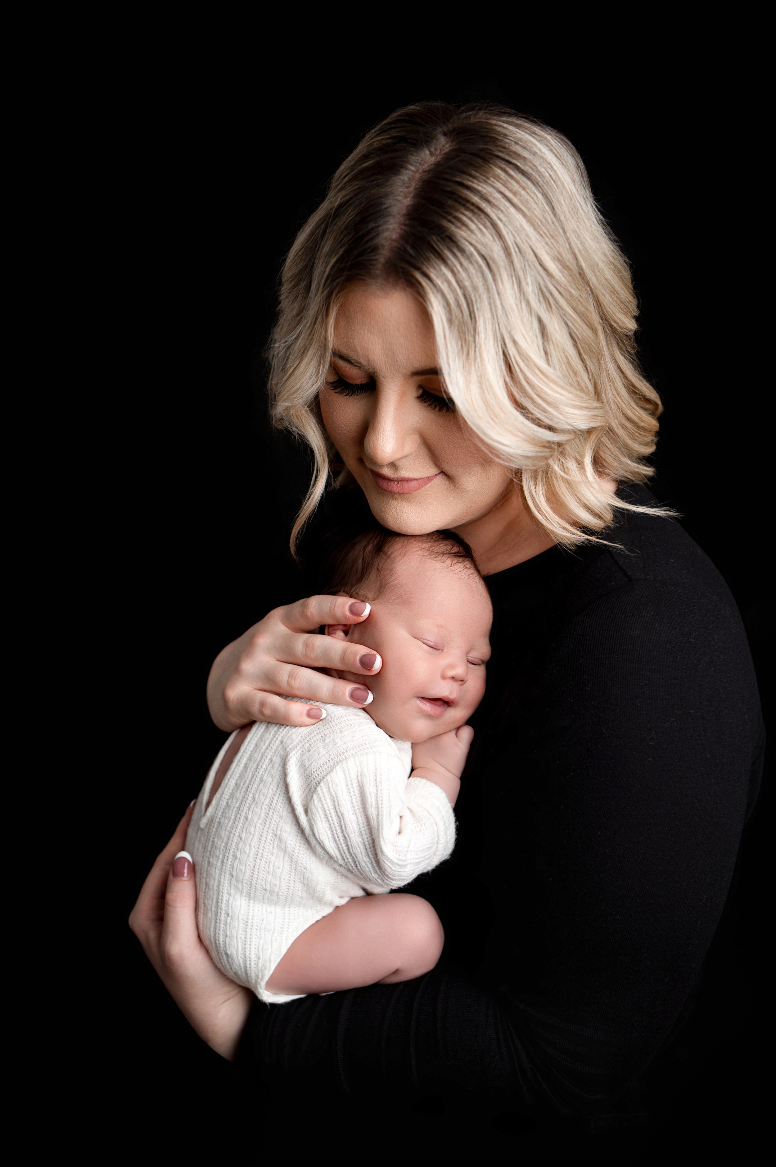 A blonde woman in a black top gently cradles a sleeping newborn baby in a white knit outfit against her chest in a dark studio portrait.