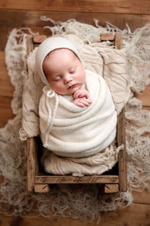 Sleeping newborn baby wrapped in cream knit blanket and bonnet, resting in a small rustic wooden crib on a textured wool rug.