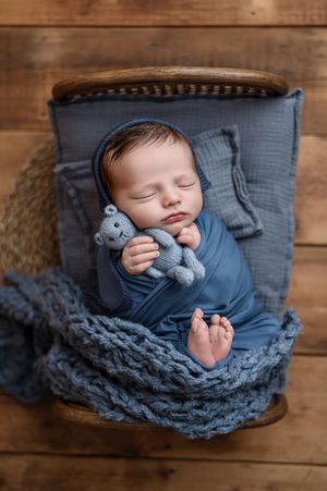 Sleeping newborn baby swaddled in blue holding a small gray knitted teddy bear on a tiny bed with a chunky knit blanket on wood.