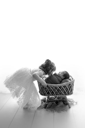 Black and white newborn photo of a young girl in a tulle dress leaning over a wicker bassinet to kiss her sleeping baby sibling.