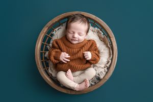 Sleeping newborn baby in a rust knit sweater and cream pants curled up in a round wooden bowl on a teal background.