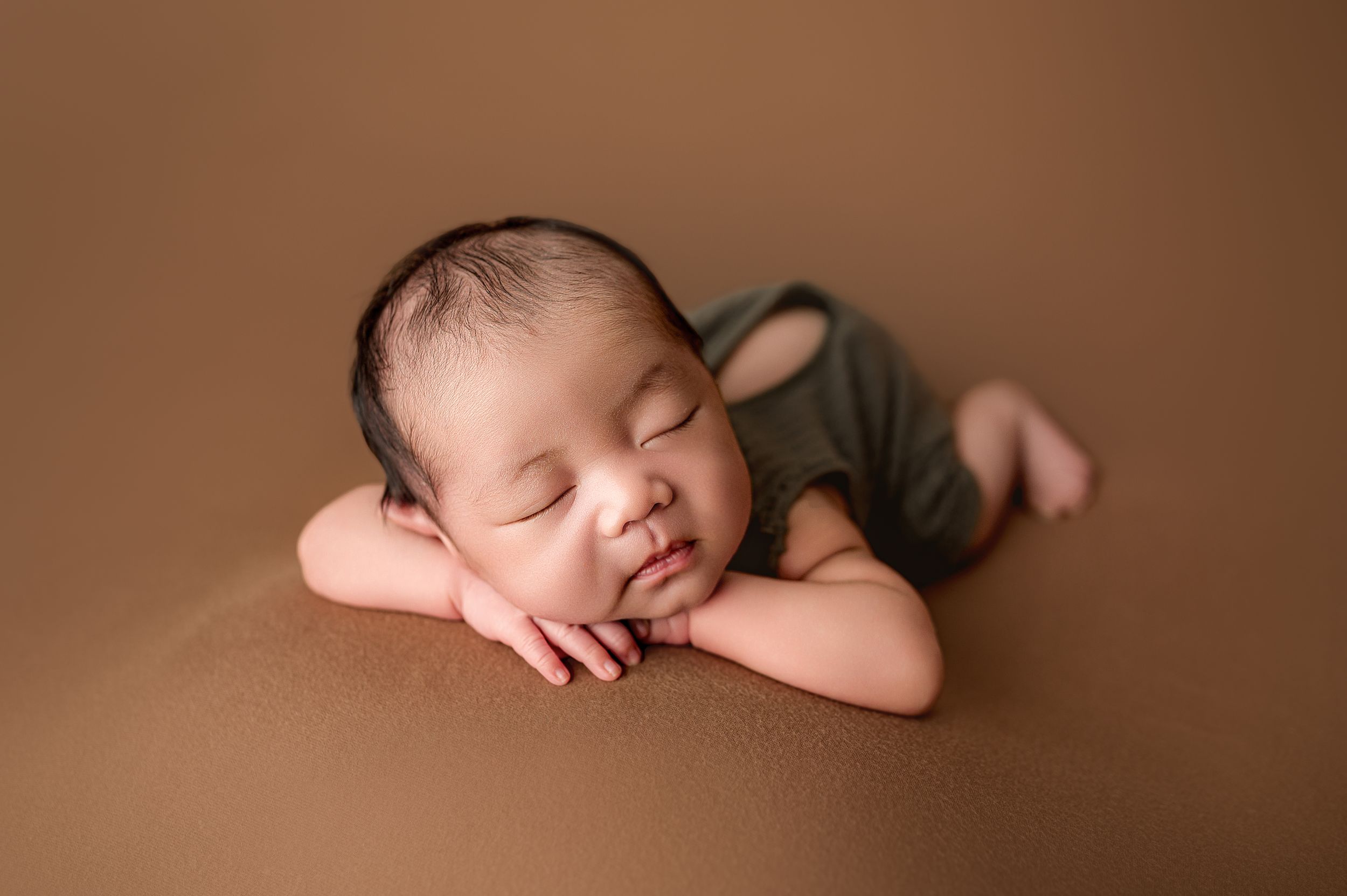 Sleeping newborn baby in an olive green romper rests head on folded arms against a warm brown backdrop in a newborn photography session.