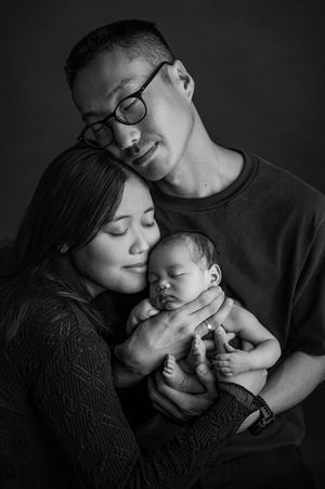 Black and white newborn portrait of parents with eyes closed, tenderly cradling their sleeping baby between them in a warm family embrace.