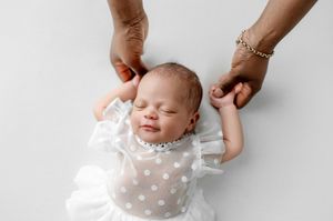 Sleeping newborn baby in a white polka dot tulle outfit, with two adult hands gently holding the infant's tiny hands on a white background.