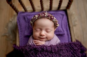 Sleeping newborn baby wearing a floral headband, wrapped in a purple knit blanket on a wooden prop bed during a studio photo session.