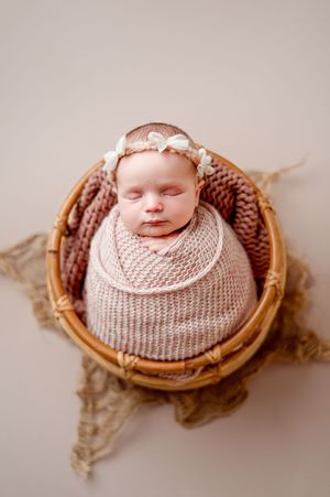 Sleeping newborn baby in a pink knit wrap and floral headband resting in a round wicker basket on a neutral backdrop.