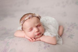 Sleeping newborn baby girl in a floral headband and white lace outfit resting her head on folded hands on a soft pastel floral blanket.