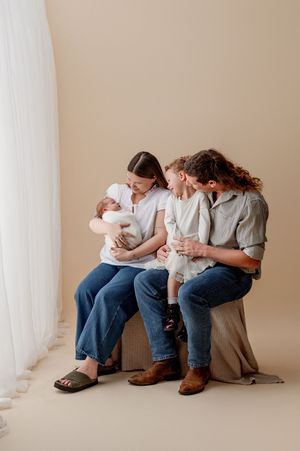 A smiling family of four sits together on a neutral studio backdrop as the mother cradles a newborn and the father holds a toddler.