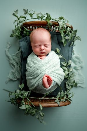 Sleeping newborn baby wrapped in a pale green knit blanket, resting in a small wicker bed decorated with leafy greenery.