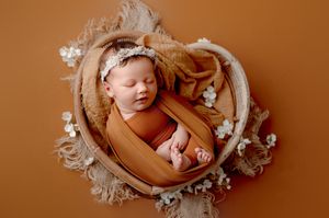 Sleeping newborn baby wrapped in a rust orange blanket in a heart-shaped basket with a floral headband and white flowers on a warm backdrop.