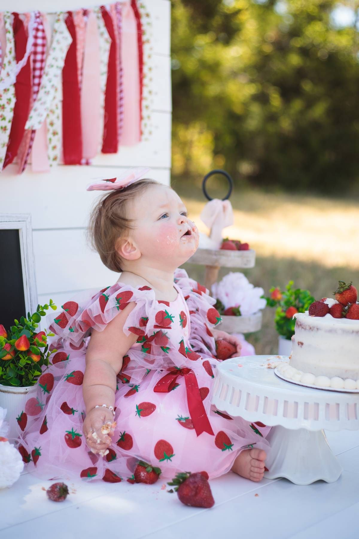 Baby girl in a pink strawberry-print dress looks up while sitting near a strawberry cake at a bright first birthday smash cake session.