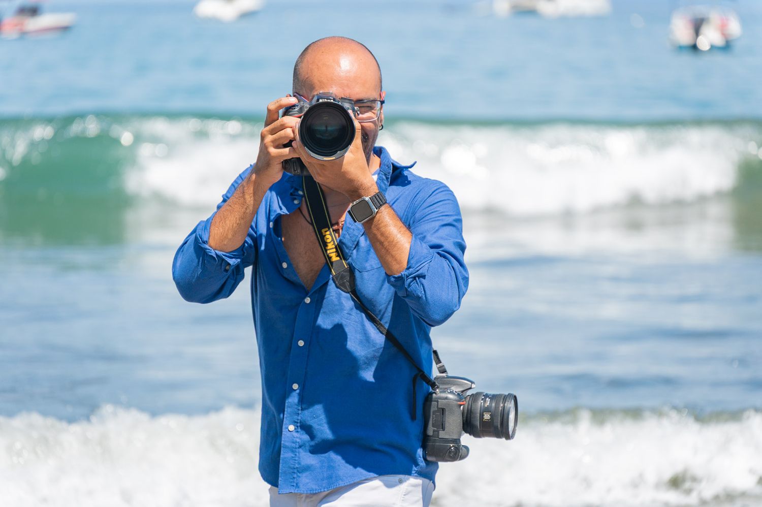 Bald man in a blue linen shirt photographs the camera at a sunny beach while ocean waves roll in and a second camera hangs from his neck.