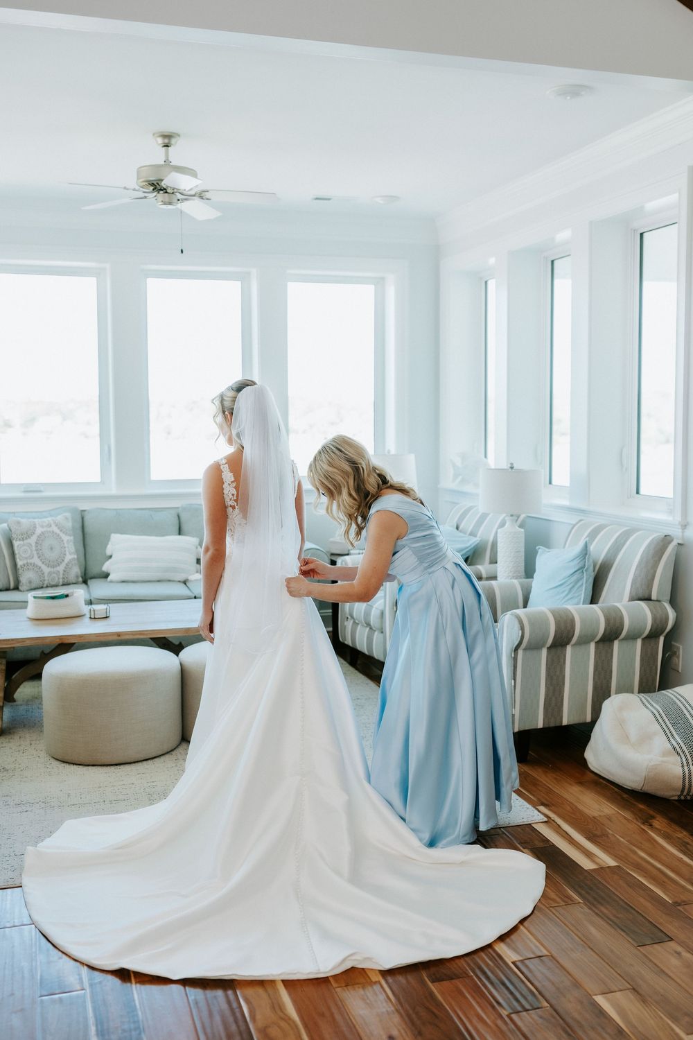 Bridesmaid in a light blue dress helps fasten a bride's white wedding gown with a long train in a bright coastal living room.