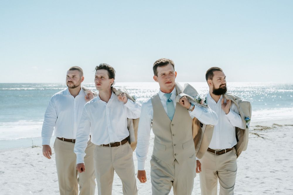 Four groomsmen in khaki pants and white shirts walk arm in arm along a sandy beach with the ocean behind them.