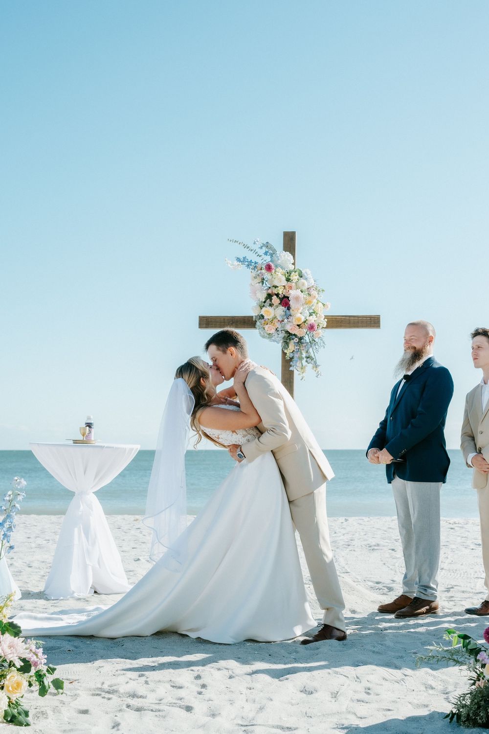 Bride and groom share a kiss during a beachfront wedding ceremony beneath a wooden cross with a floral arrangement as guests look on.
