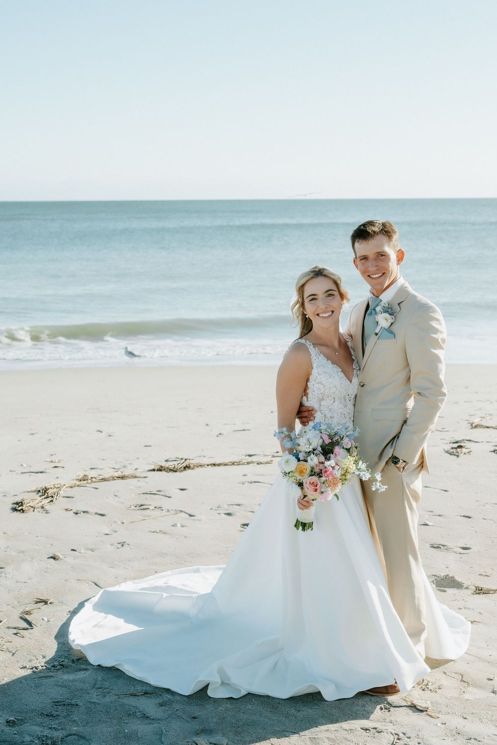 Smiling bride in a white gown holding a pastel bouquet poses with her groom in a tan suit on a sandy beach by the ocean.