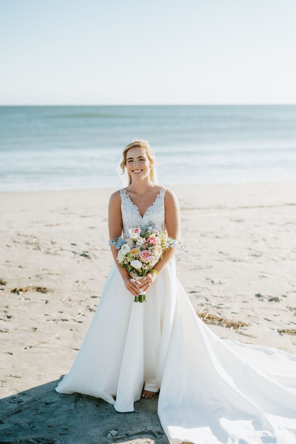 Smiling bride in a white lace-bodice gown with a long train holds a pink and white bouquet while standing barefoot on a sunny beach.