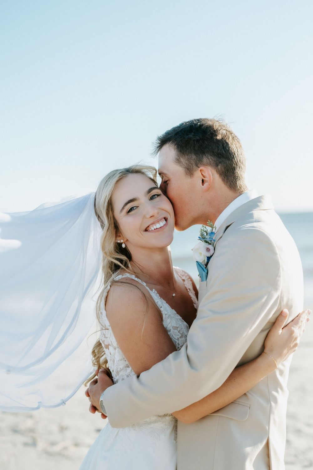 Smiling bride in a lace wedding dress with her veil blowing in the wind embraces a groom in a tan suit kissing her cheek on a sunny beach.