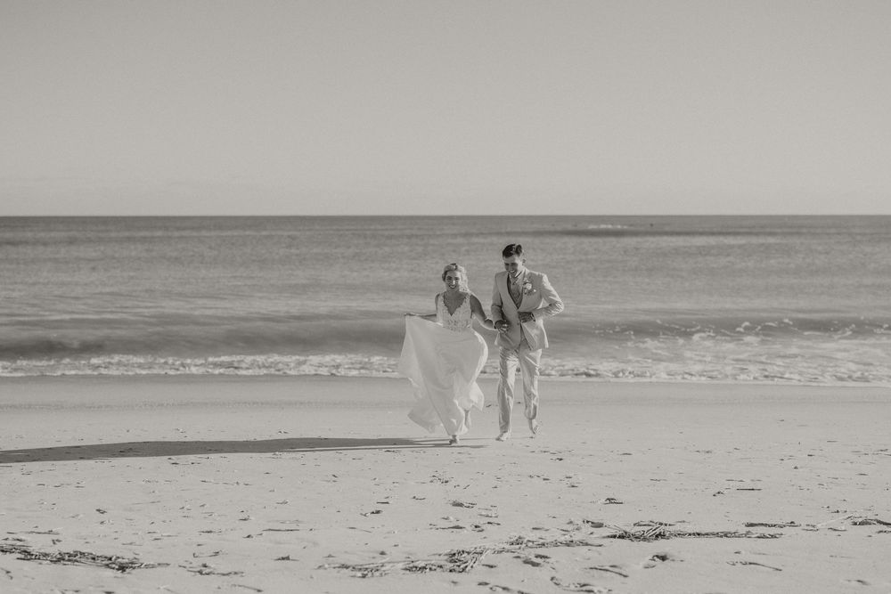 Black and white photo of a bride and groom walking hand in hand along a sandy beach with ocean waves behind them.
