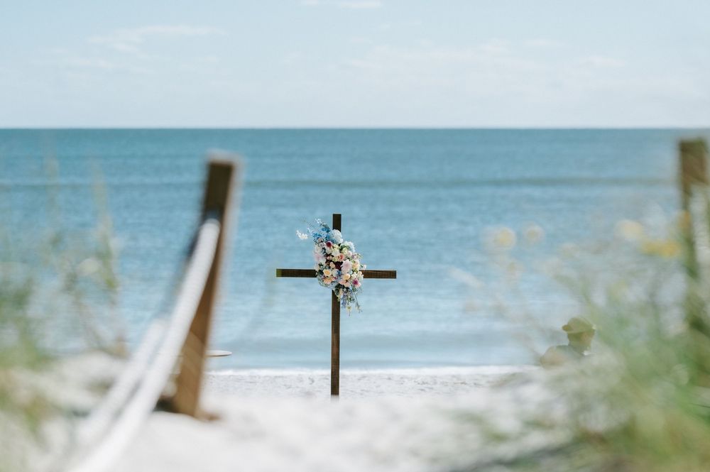 Wooden cross decorated with white flowers stands on a sandy beach path overlooking calm blue ocean waters under a clear sky.