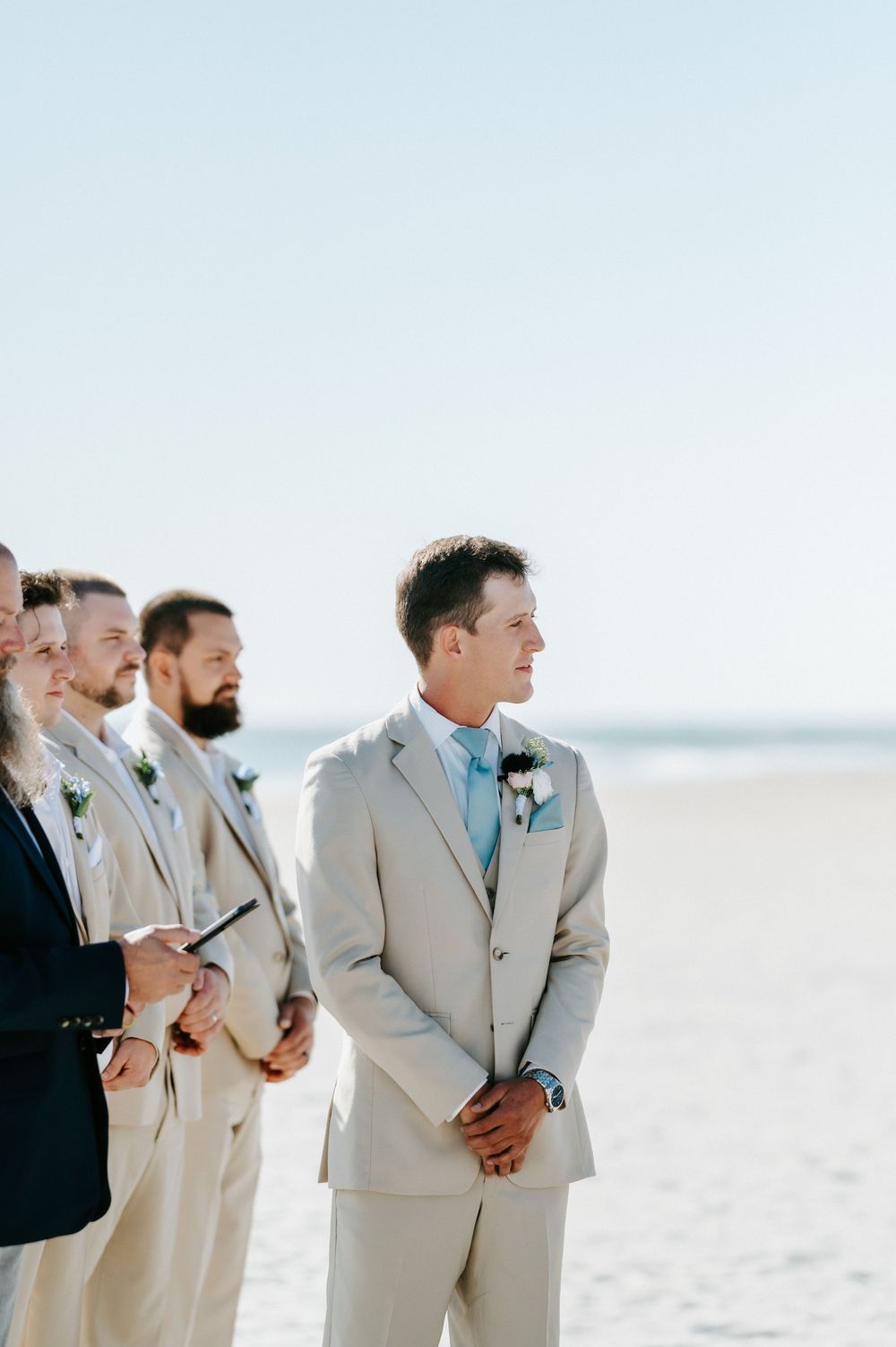 Groom in a tan suit and light blue tie stands smiling on a sunny beach, with groomsmen in matching suits beside him at a wedding ceremony.