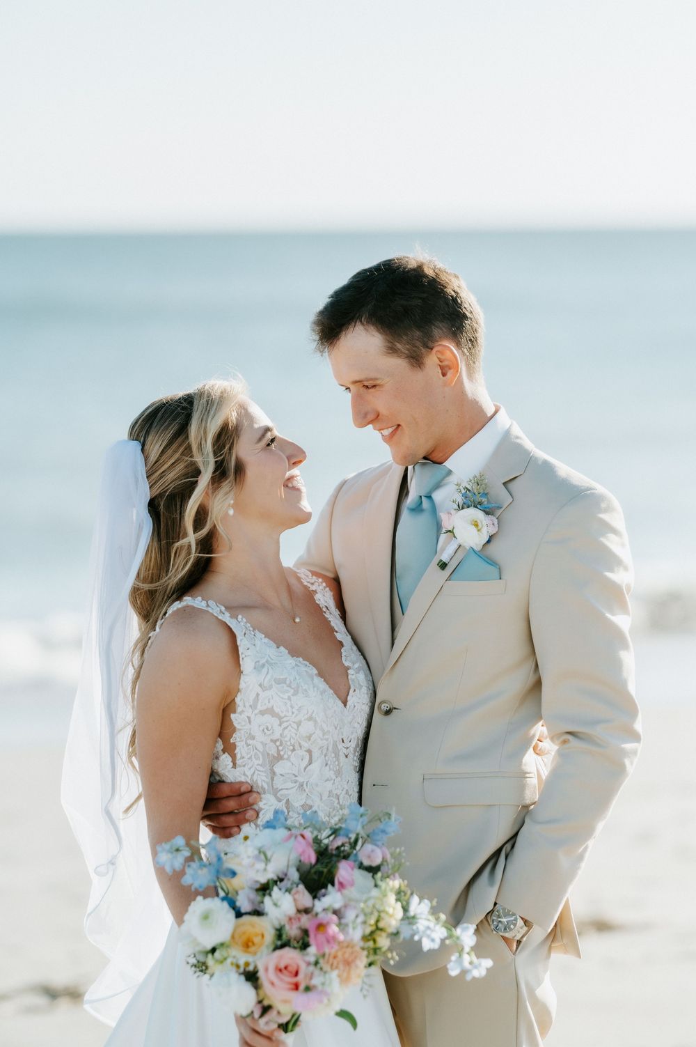 Smiling bride in a lace gown and veil embraces her groom in a tan suit on a sunny beach, holding a pastel bouquet.