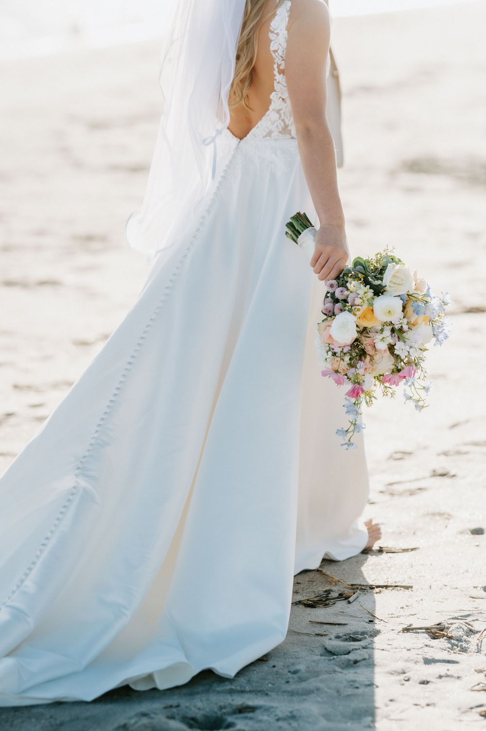 Bride in a flowing white gown with a lace back and veil holds a pastel wildflower bouquet while standing barefoot on a sandy beach.