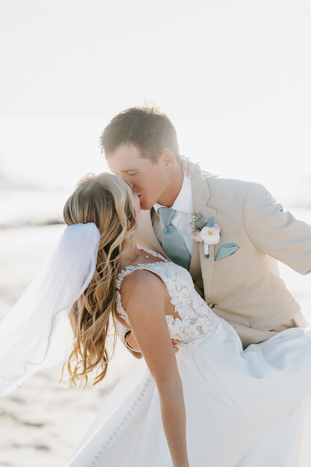 Bride in a lace wedding gown and long veil kisses groom in a tan suit with a floral boutonniere during a beach wedding.
