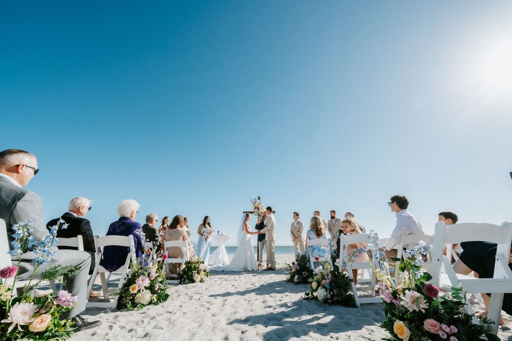 Beach wedding ceremony on white sand under a clear blue sky, with guests seated in white chairs beside colorful floral arrangements.