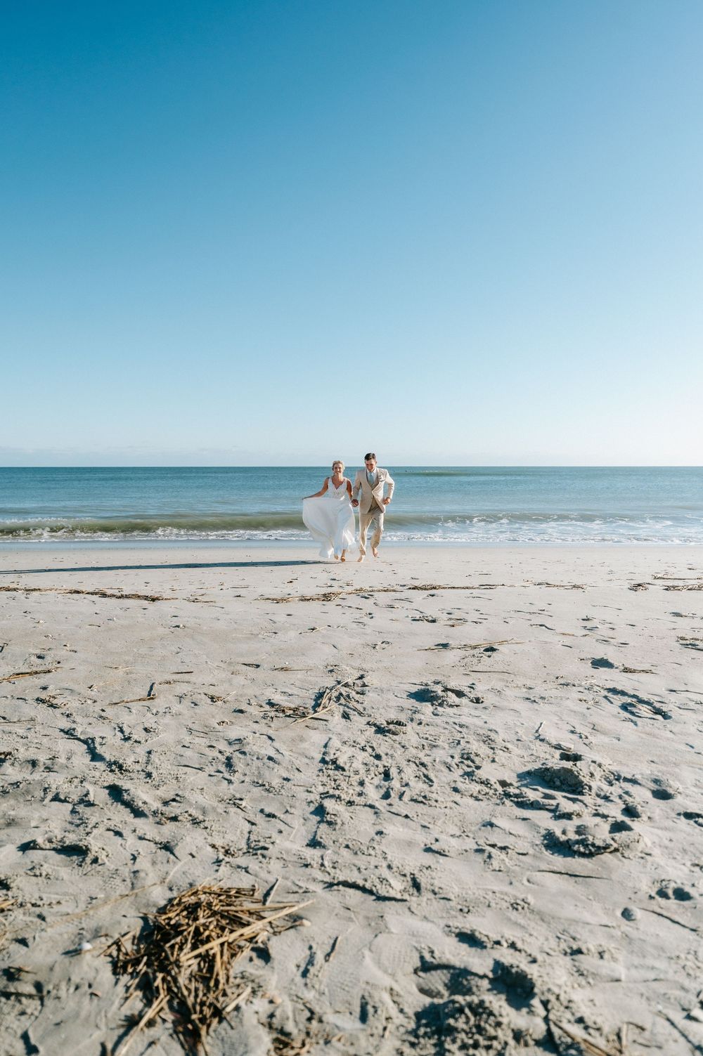 Bride and groom walk hand in hand along a wide sandy beach toward the ocean under a clear blue sky, a seaweed pile in the foreground.