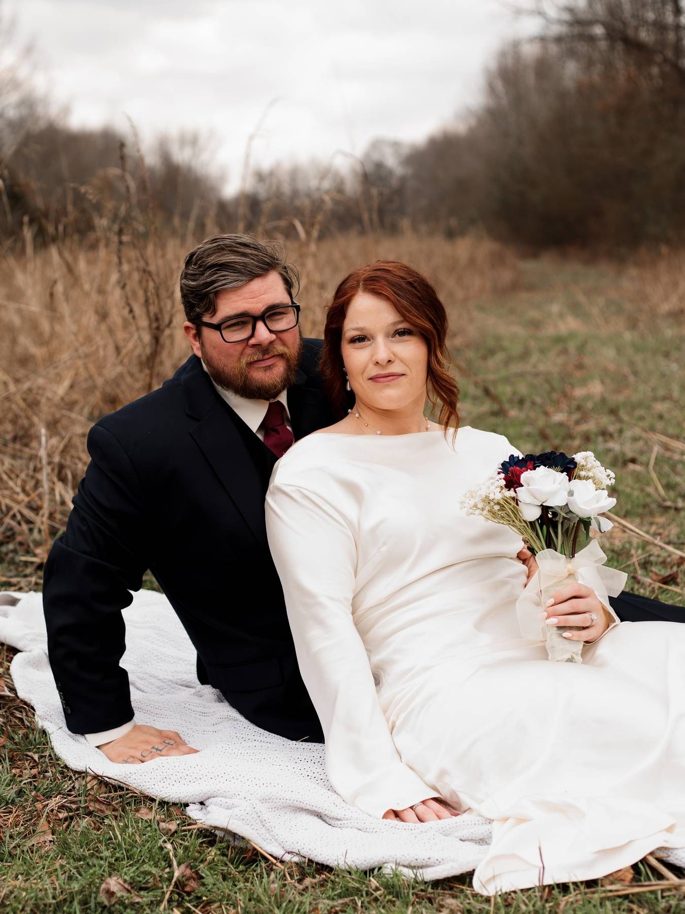 A bride in a long-sleeved white gown and a bearded groom in a dark suit and glasses sit on a blanket in an autumn field.