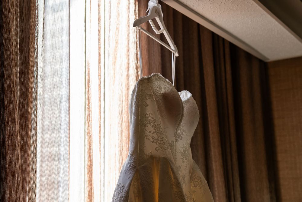 A strapless ivory lace wedding gown hangs on a white hanger beside a sunlit window with warm brown curtains.