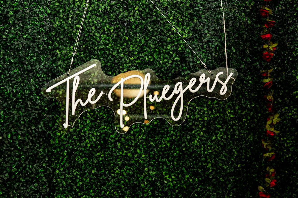 White neon sign reading The Pluegers in cursive script hanging against a lush green boxwood hedge wall, perfect for wedding decor.