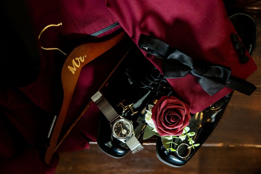 Groom's wedding morning flat lay with a burgundy suit jacket, black bow tie, silver watch, red rose boutonniere and a wooden Mr hanger.