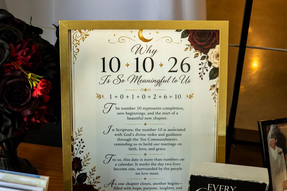 Framed wedding sign titled "Why 10-10-26 Is So Meaningful to Us" with gold border, floral accents, and dark red roses on a reception table.