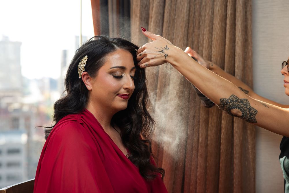 A bride in a red dress sits by a sheer curtained window while a makeup artist applies setting spray to her styled dark hair.