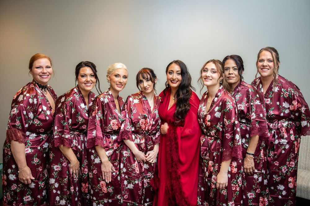Bride in a red satin robe stands among eight smiling bridesmaids wearing matching burgundy floral robes while getting ready for the wedding.