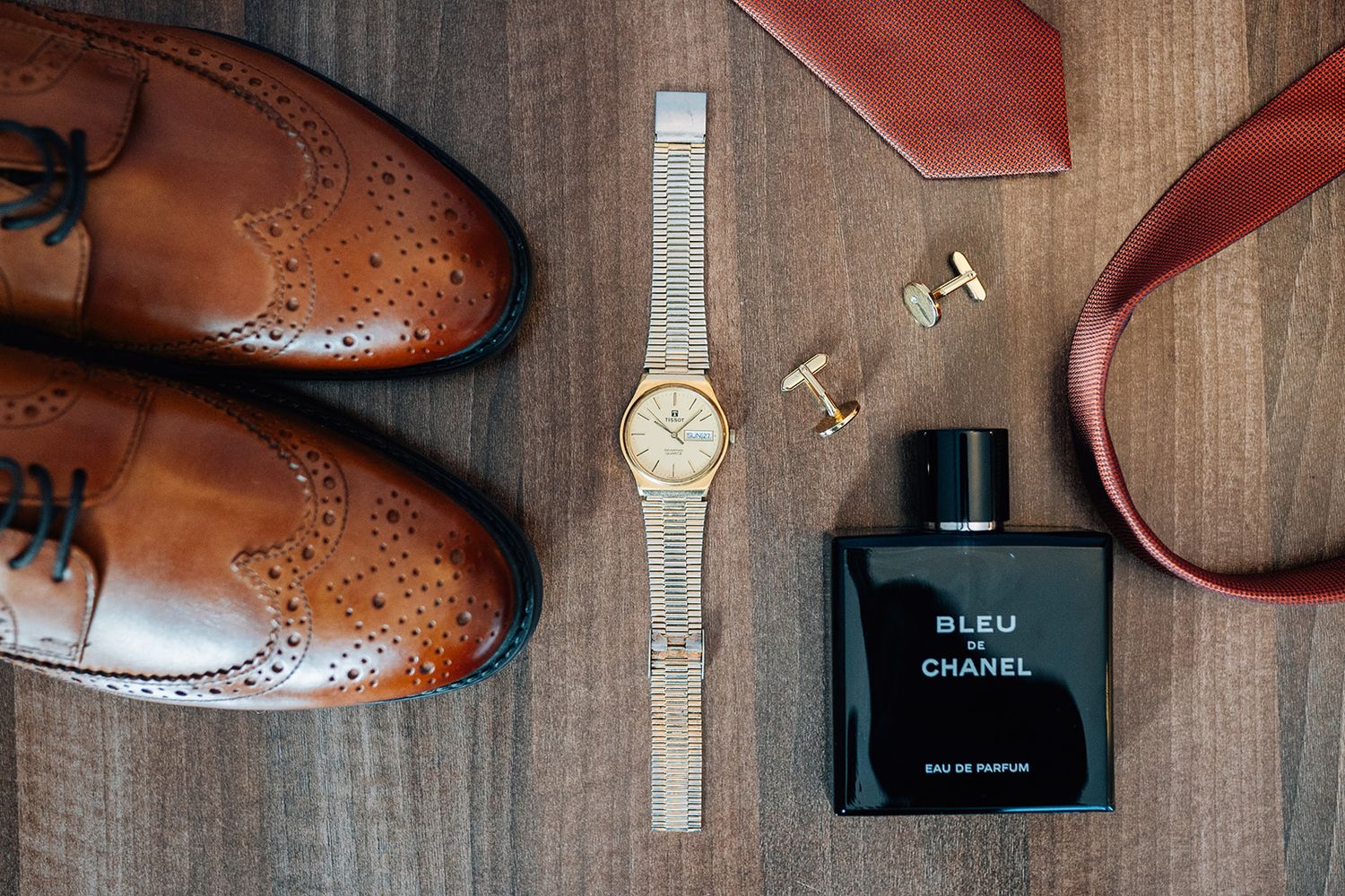 Brown leather brogue shoes, gold wristwatch, cufflinks, Bleu de Chanel cologne and a red tie arranged on a wooden surface.