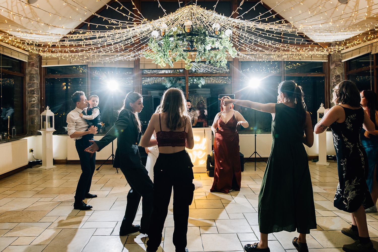 Guests in formal attire dance on a tiled floor at an evening wedding reception under string lights and a floral chandelier.