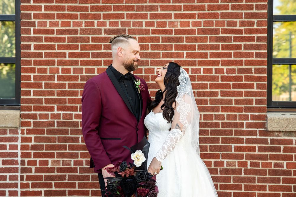 Bride in a white lace gown and veil laughs with her bearded groom in a burgundy blazer, holding a bouquet, against a red brick wall.