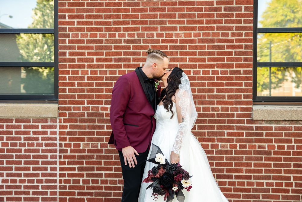 Bride in a white lace gown holding a dark floral bouquet kisses groom in a burgundy blazer in front of a red brick building.