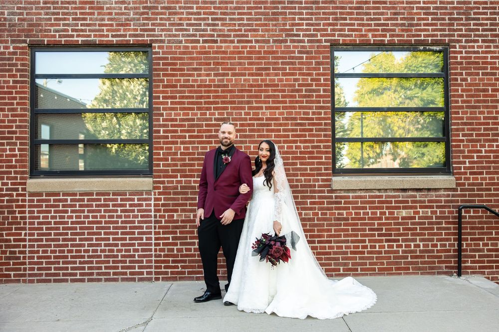 Bride in a white gown holding a burgundy bouquet poses beside a groom in a burgundy blazer in front of a brick wall with large windows.