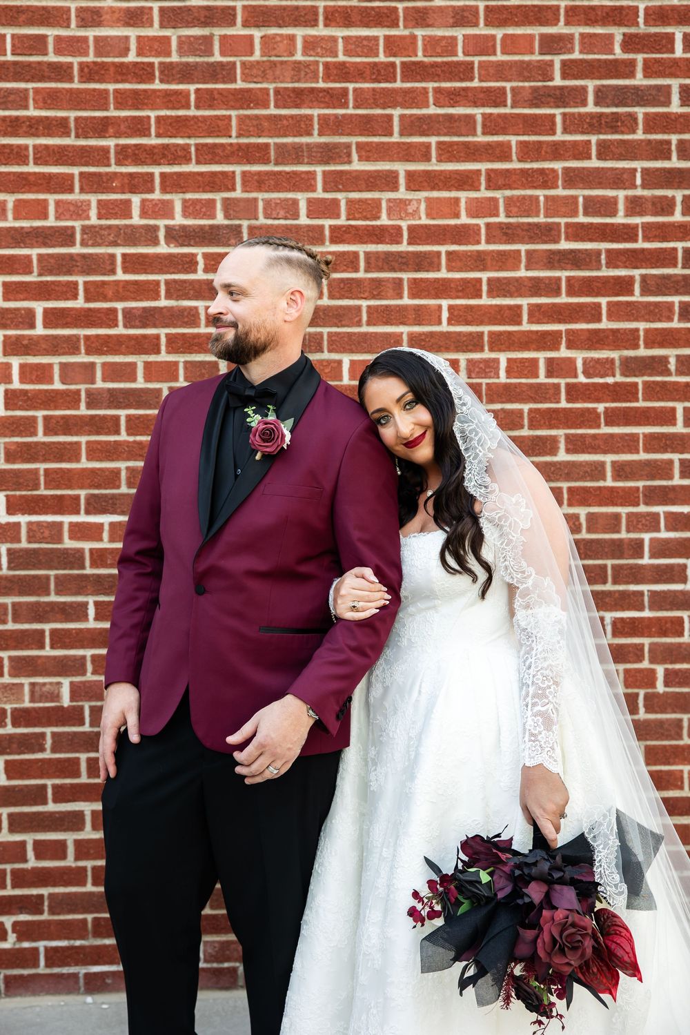 Bride in a lace gown and veil leans on a groom in a burgundy tuxedo jacket, holding a dark floral bouquet against a red brick wall.