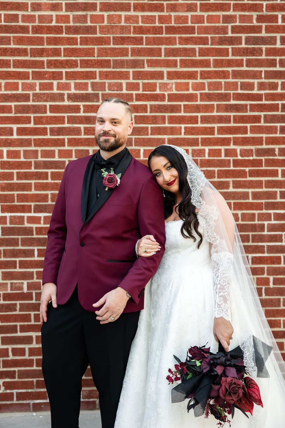Bride in a lace gown and veil holding a dark red bouquet stands arm in arm with a groom in a burgundy tuxedo jacket by a brick wall.