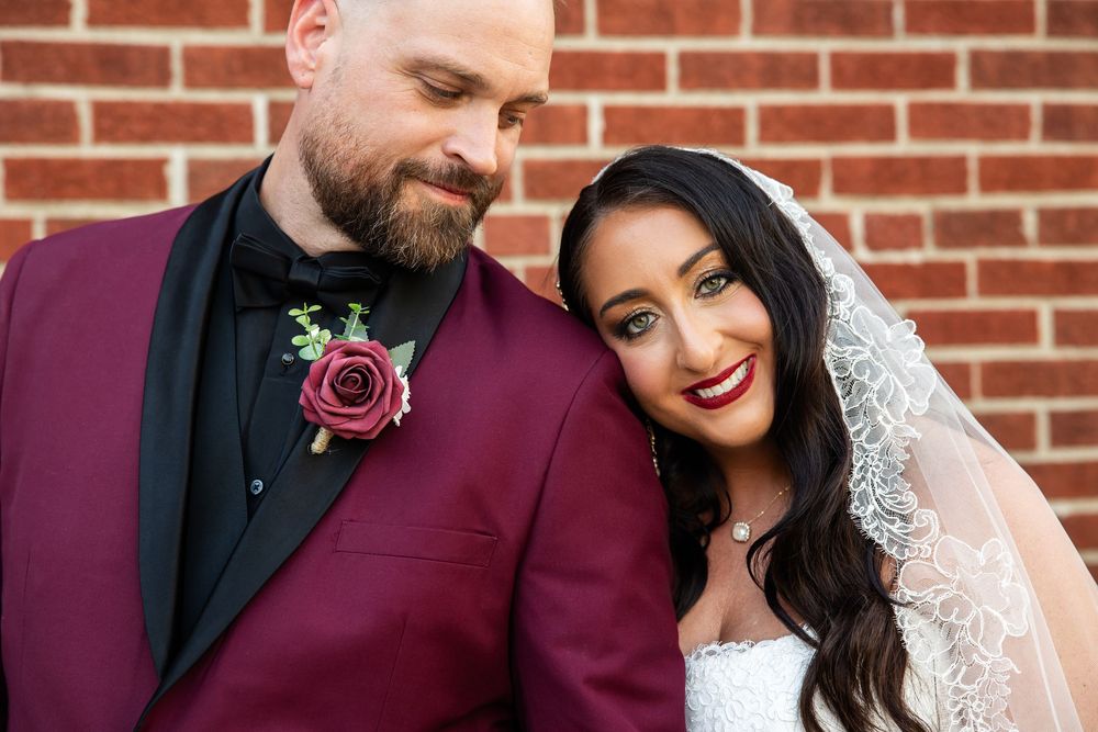 Smiling bride in a lace veil leans her head against a bearded groom in a burgundy tuxedo jacket with a rose boutonniere by a brick wall.