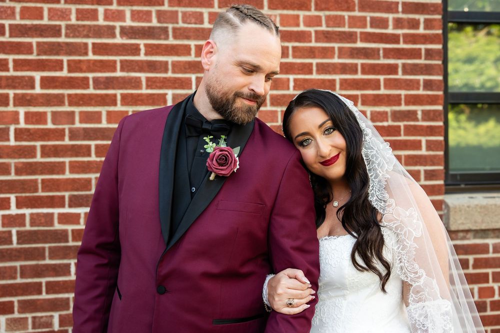Smiling bride in a white gown and lace veil leans her head on the shoulder of a bearded groom in a burgundy suit with a rose boutonniere.