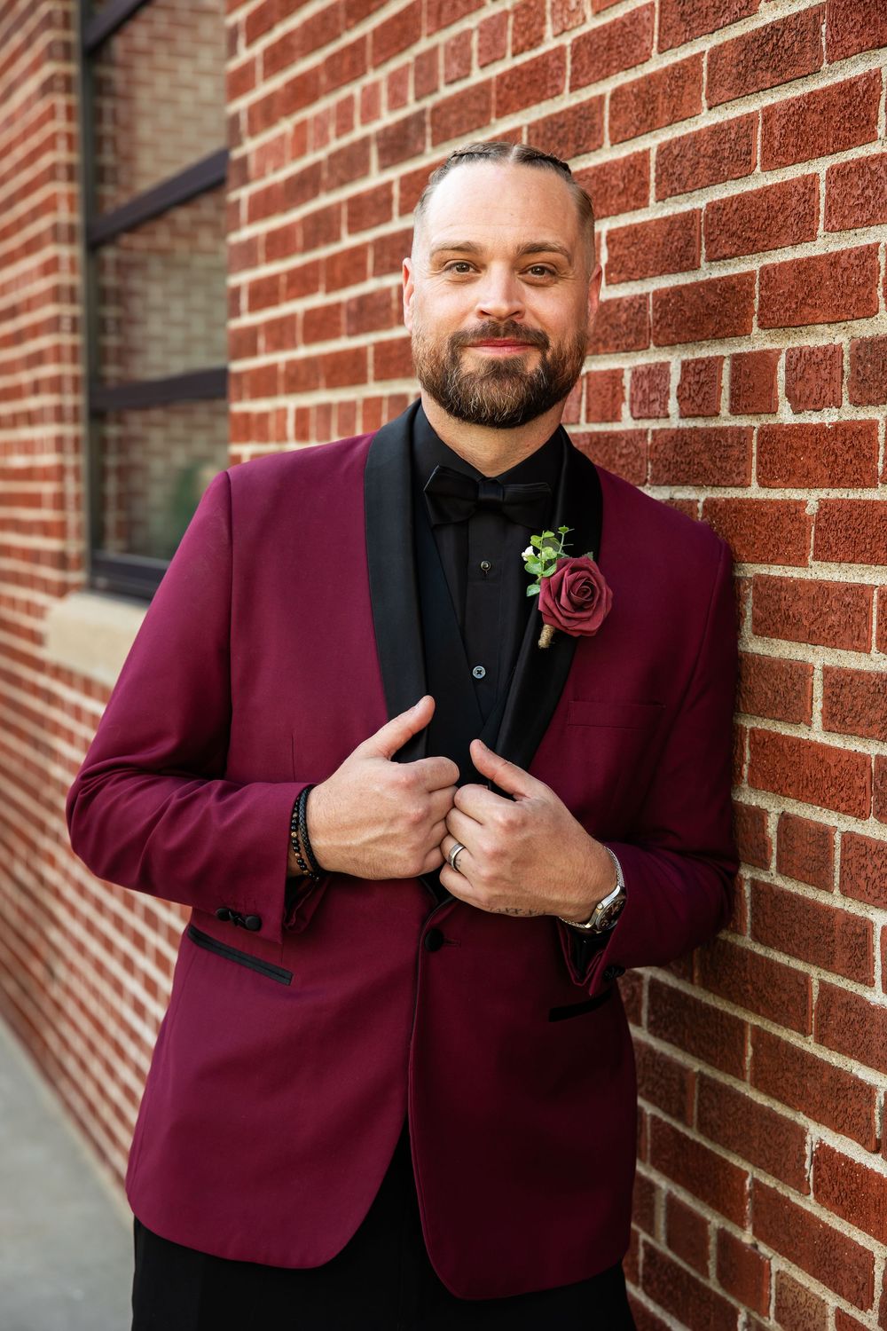 Bearded groom in a burgundy tuxedo jacket with black lapels and a floral boutonniere leaning against a red brick wall.