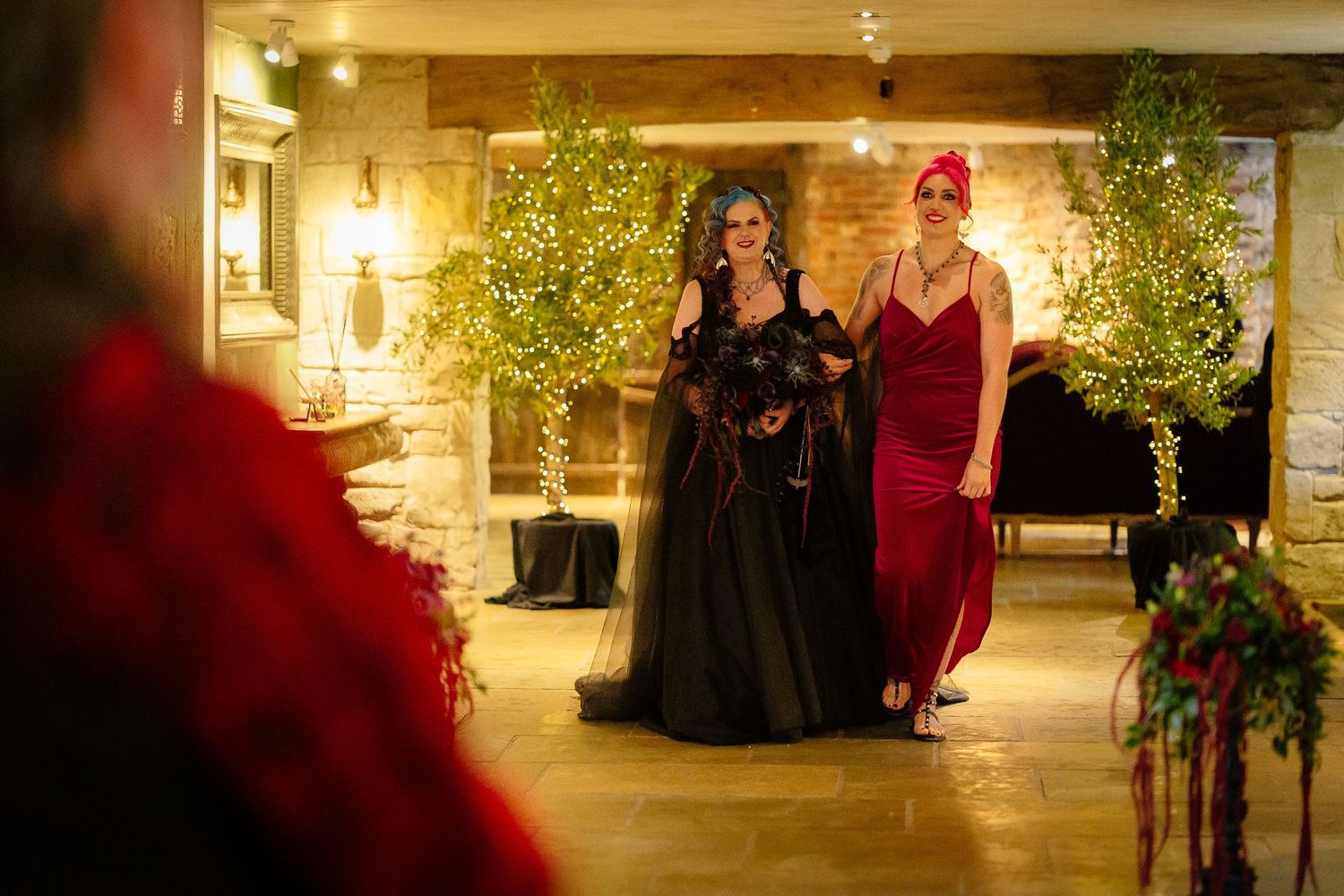 Bride in a black wedding dress walking in with her bridesmaid between fairy-lit trees at Le Petit Chateau